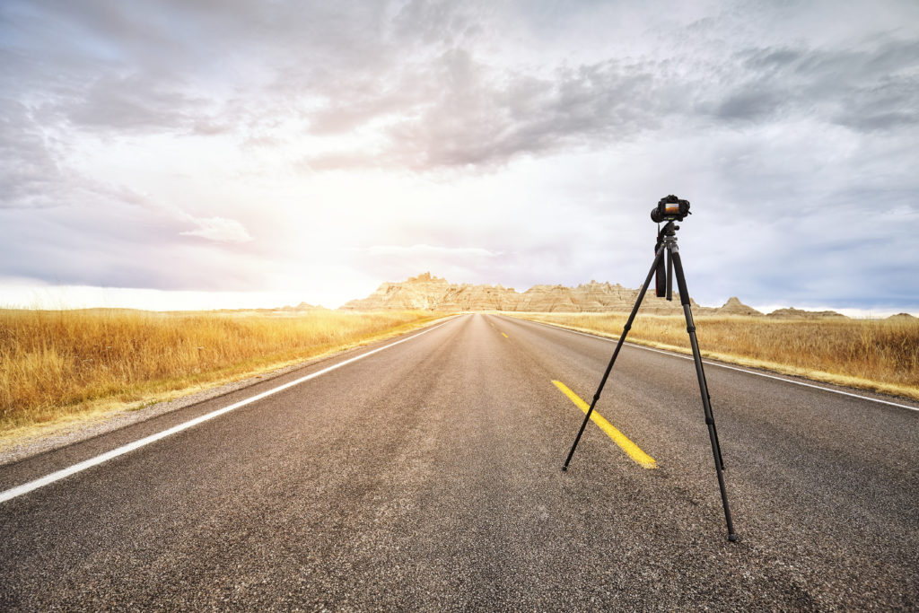 Professional photo camera on tripod on an empty road at sunset, focus on the camera, travel or work concept, Badlands National Park, South Dakota, USA.